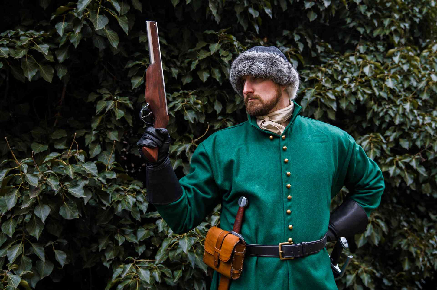 Medieval reenactor in green coat and fur hat holding raised wheellock blaster pistol outdoor LARP