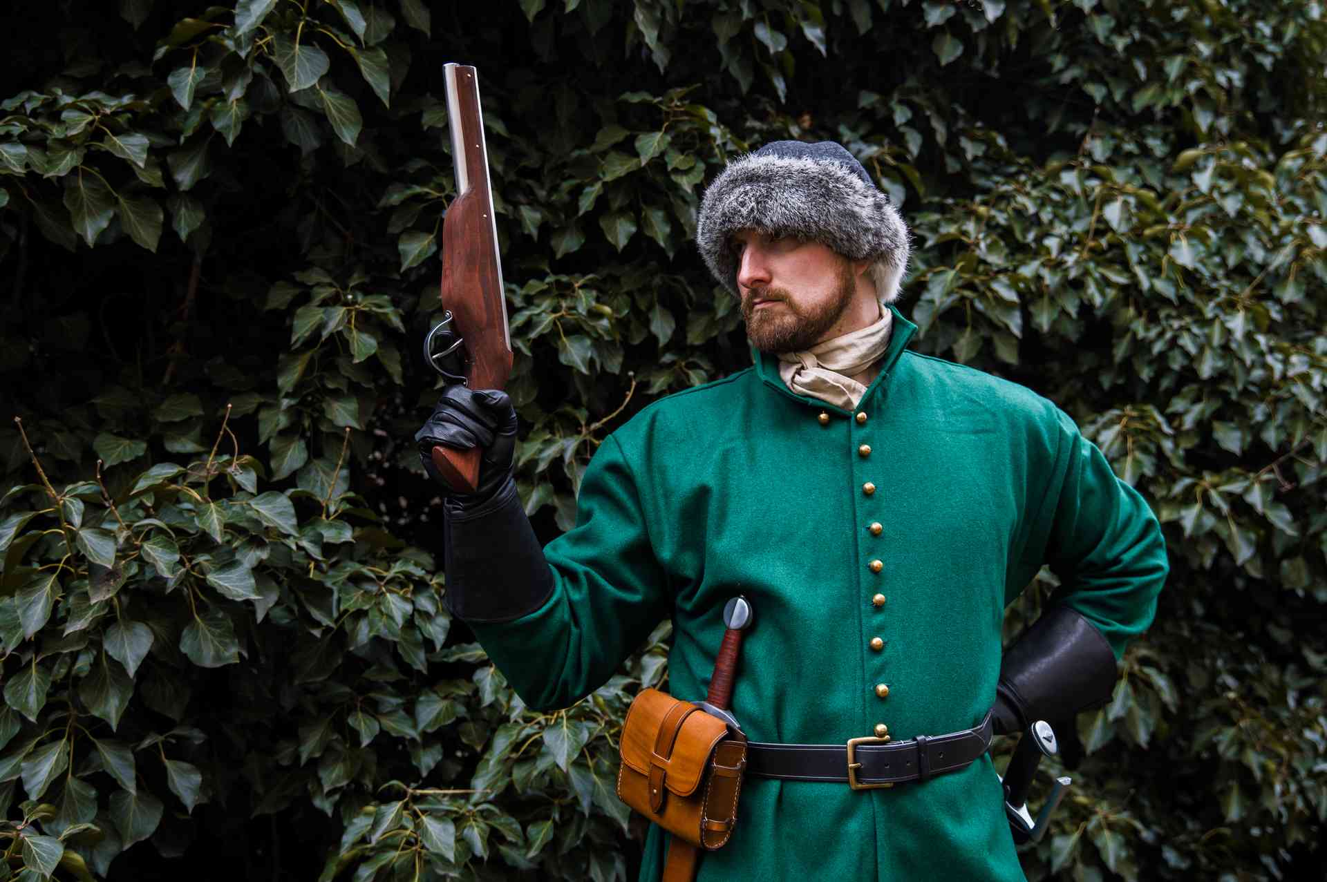 Medieval reenactor in green coat and fur hat holding raised wheellock blaster pistol outdoor LARP