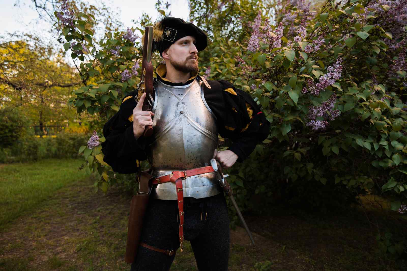 Medieval soldier in armor holding musket in lilac garden with purple flowers LARP weapon