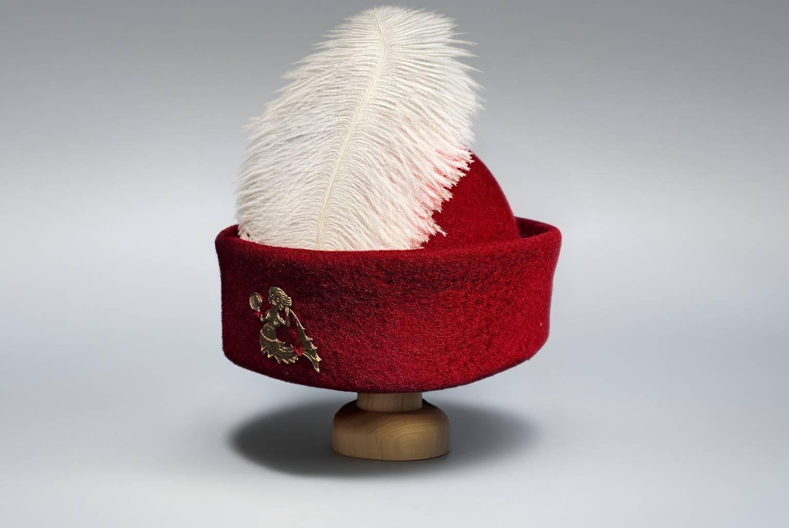Red round cap wool felt hat with white feather and golden brooch on wooden stand